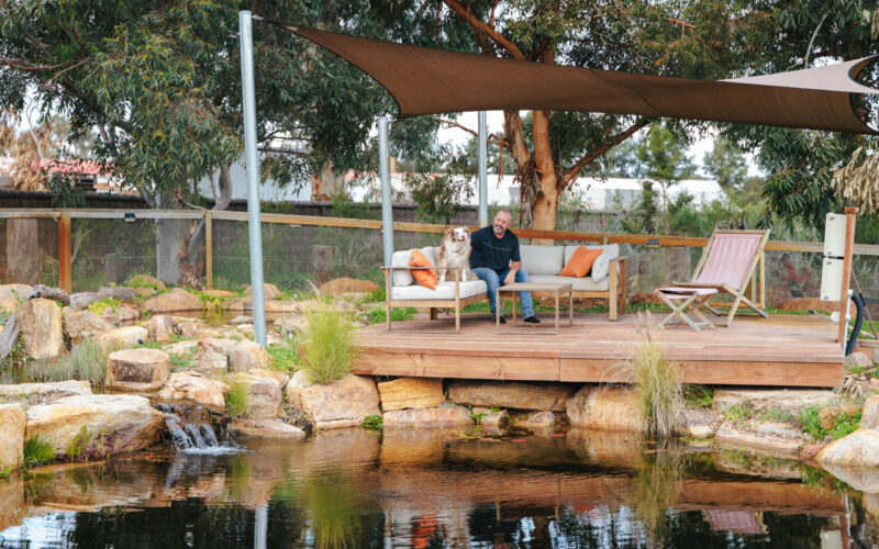 Client seated on a timber deck beside a landscaped dam with shade sail